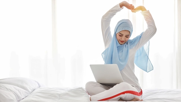 Mujer árabe asiática joven que usa la computadora portátil que se sienta en la cama en el dormitorio. Hermosa chica musulmana en ropa de dormir con hijab azul estirando sus brazos después de trabajar antes de irse a la cama. Concepto de estilo de vida y tecnología