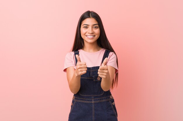 La mujer árabe bastante joven que lleva un mono de los vaqueros con los pulgares sube, anima sobre algo, apoya y respeta el concepto.