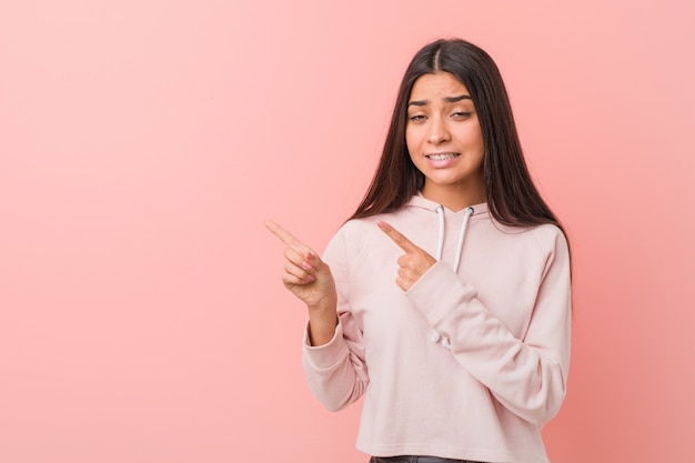 La mujer árabe bonita joven que lleva un deporte casual parece sorprendido señalando con los dedos índices a un espacio de la copia.