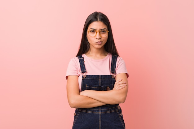 La mujer árabe bonita joven que lleva un mono que frunce el ceño cara de los vaqueros en disgusto, mantiene los brazos cruzados.