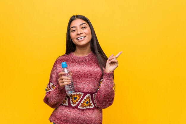 Mujer árabe joven que sostiene una botella de agua que sonríe alegremente que señala con el índice lejos.