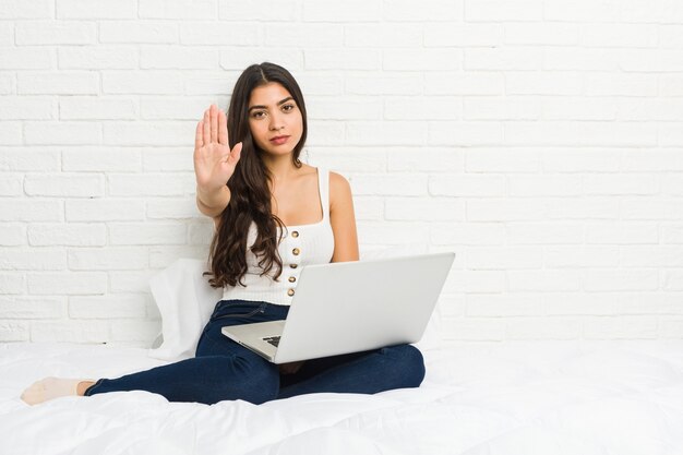 Mujer árabe joven que trabaja con su computadora portátil en la cama que se coloca con la mano extendida que muestra la señal de stop, previniéndole.