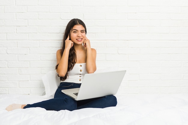 Mujer árabe joven que trabaja con su computadora portátil en la cama que cubre los oídos con las manos.