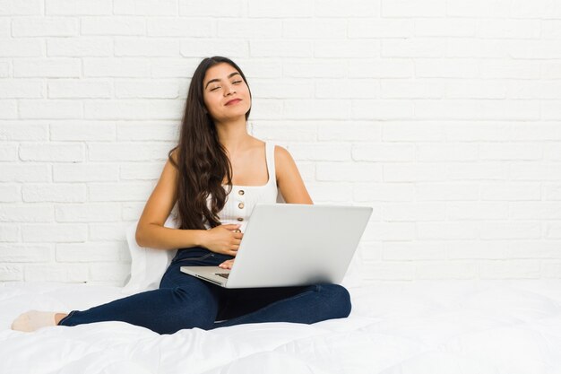 La mujer árabe joven que trabaja con su computadora portátil en la cama toca la panza, sonríe suavemente, comiendo y concepto de la satisfacción.