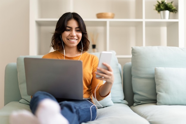 Mujer árabe joven sonriente que usa la computadora portátil y el teléfono inteligente mientras descansa en casa