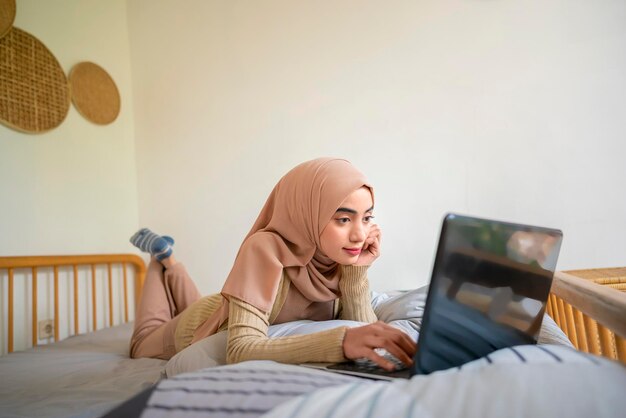 Una mujer árabe musulmana está acostada en una cama con una computadora portátil en el regazo