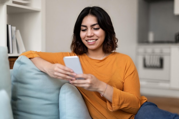 Mujer árabe sonriente usando un teléfono inteligente mientras se relaja en el sofá en casa