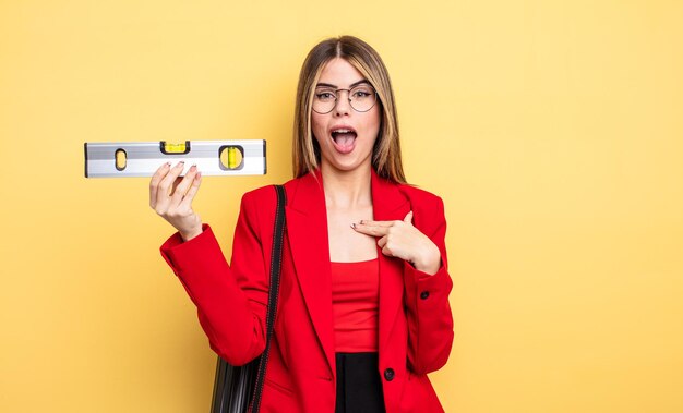 Mujer arquitecta que parece sorprendida y sorprendida con la boca abierta, apuntándose a sí misma y manteniendo un equilibrio nivelado