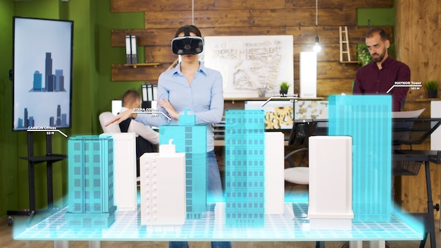 Foto la mujer del arquitecto con casco de realidad virtual está mirando proyecciones holográficas de la construcción de una ciudad. ella reorganiza los pisos de una torre comercial