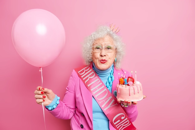 Mujer arrugada senior de pelo rizado envejecido mantiene los labios redondeados sostiene un delicioso pastel con velas encendidas El globo inflado celebra el 91 cumpleaños viste ropa festiva