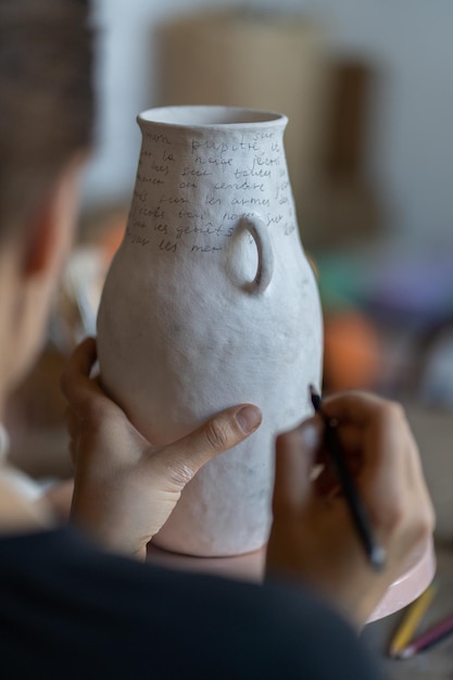 Mujer artesana pinta texto en jarrón de cerámica blanca con marcador sobre fondo borroso en taller