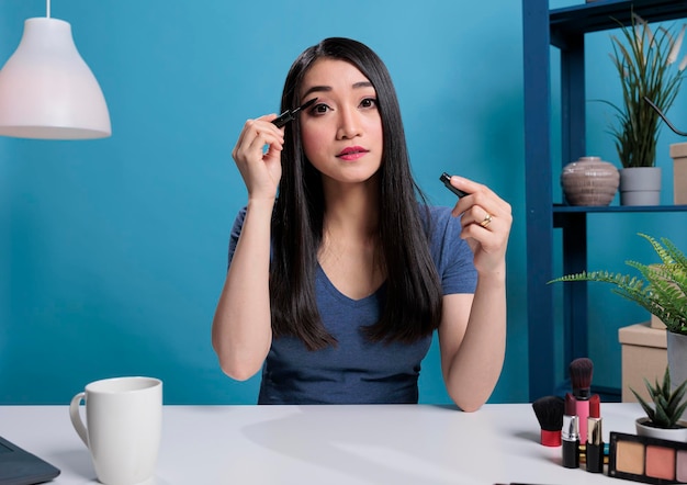 Mujer artista asiática grabando tutorial de maquillaje usando rímel negro publicitando productos cosméticos en estudio con fondo azul. Influenciador de las redes sociales filmando revisión de maquillaje para contenido de podcast