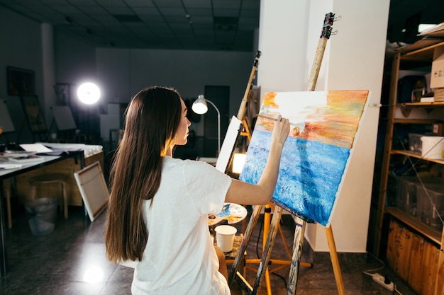 Mujer artista pintando un cuadro en caballete con pinturas al óleo en su taller