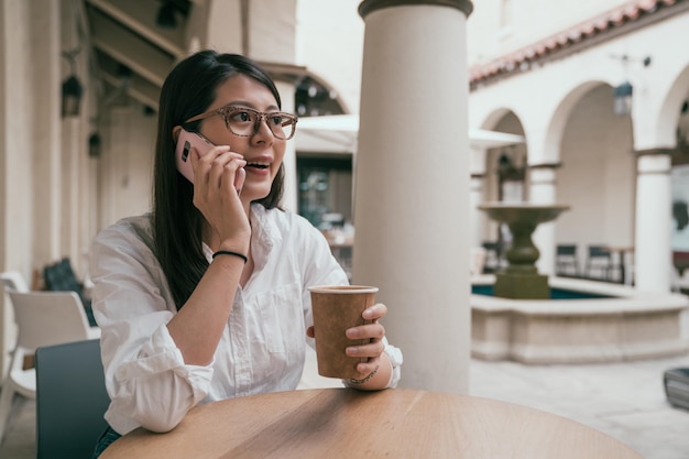 mujer asaina hablando por teléfono y sentada en el café bar con una taza de café en la mano.