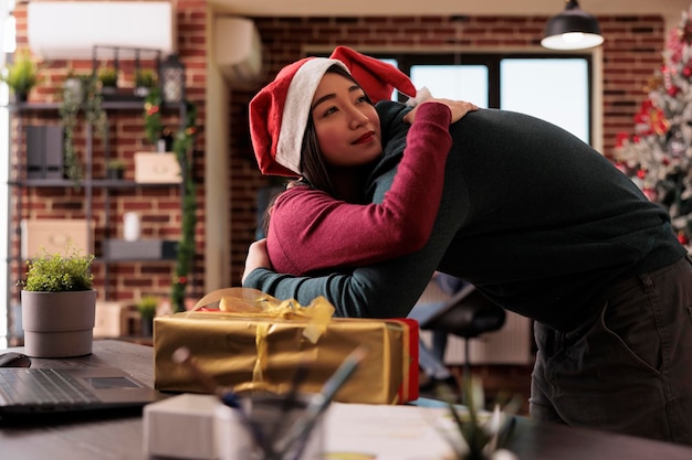 Mujer asiática abrazando a un compañero de trabajo después de recibir un regalo en la oficina de inicio decorada con adornos navideños festivos. Celebrando la temporada de vacaciones de invierno con árbol de Navidad y caja de regalo, tradición.
