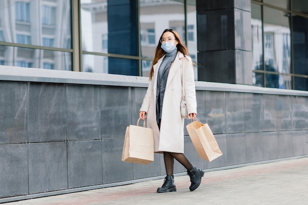 Mujer asiática con un abrigo, gafas y una máscara médica se encuentra en la calle, sosteniendo un paquete en sus manos.