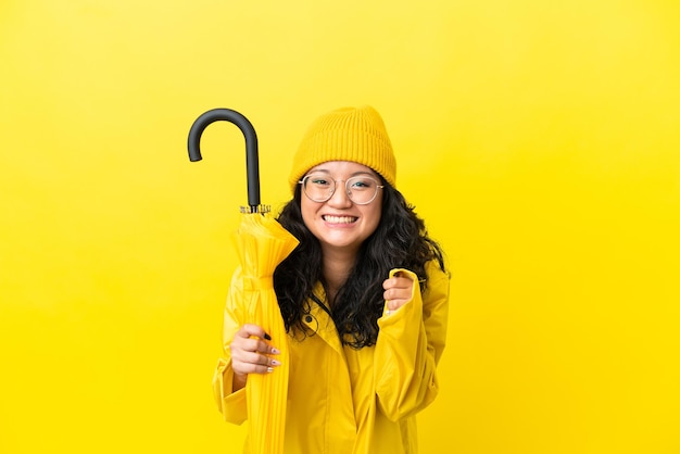 Mujer asiática con abrigo impermeable y paraguas aislado sobre fondo amarillo celebrando una victoria en la posición ganadora