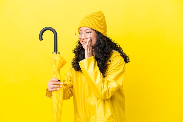 Foto mujer asiática con abrigo impermeable y paraguas aislado sobre fondo amarillo gritando con la boca abierta hacia un lado