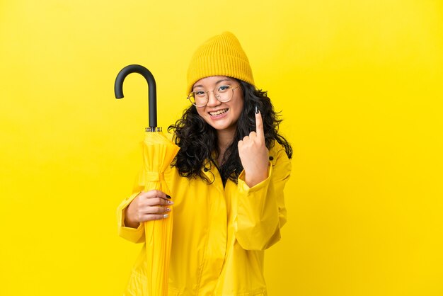 Mujer asiática con abrigo impermeable y paraguas aislado sobre fondo amarillo haciendo gesto que viene