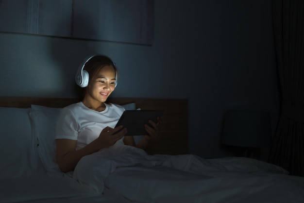 Mujer asiática adicta al móvil que usa tableta digital navegando por Internet para ver películas o videollamadas de reuniones virtuales en línea con su novio en la cama antes de dormir por la noche.