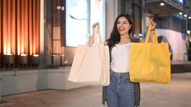 Mujer asiática alegre que lleva bolsas de compras y camina por la calle de la ciudad por la noche