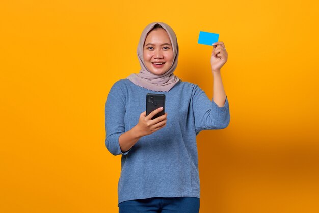 Mujer asiática alegre que sostiene el teléfono móvil y que muestra la tarjeta de crédito sobre fondo amarillo