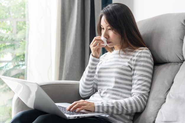 Mujer asiática alérgica que sopla la nariz en un pañuelo de papel sentarse en el sofá en casa, estudiar o trabajar en una computadora portátil, la niña enferma se resfrió con síntomas de alergia sosteniendo una servilleta en la mano mientras trabajaba en la computadora