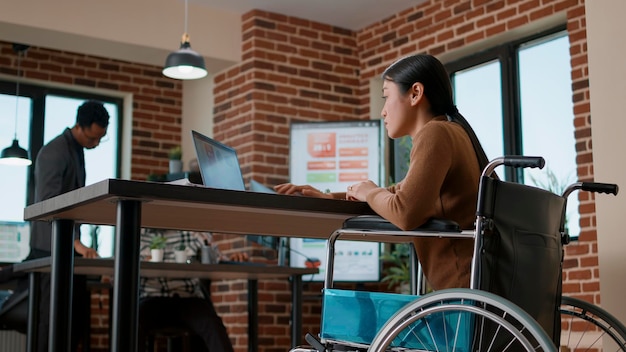 Mujer asiática analizando gráficos financieros en computadoras portátiles, lidiando con discapacidad crónica. Trabajador de oficina mirando gráficos en la pantalla del dispositivo, planeando marketing y estrategia de liderazgo.