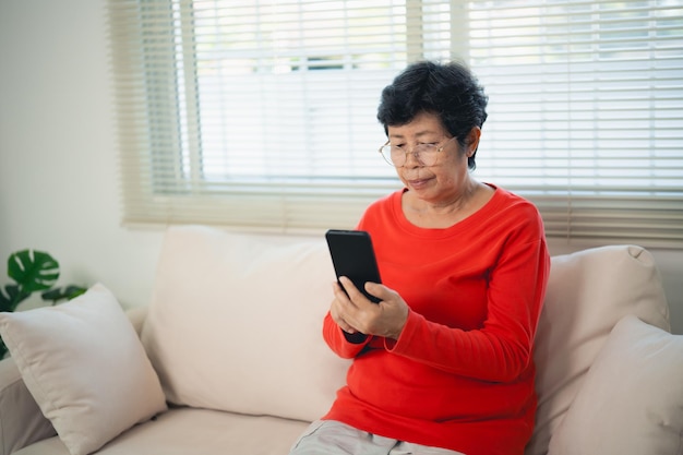 Mujer asiática anciana feliz disfrutando de usar aplicaciones móviles enviando mensajes de texto escribiendo mensajes sentada en el sofá sonriendo anciana con teléfono inteligente mirando teléfono inteligente navegando por las redes sociales o aprendiendo tecnología en casa