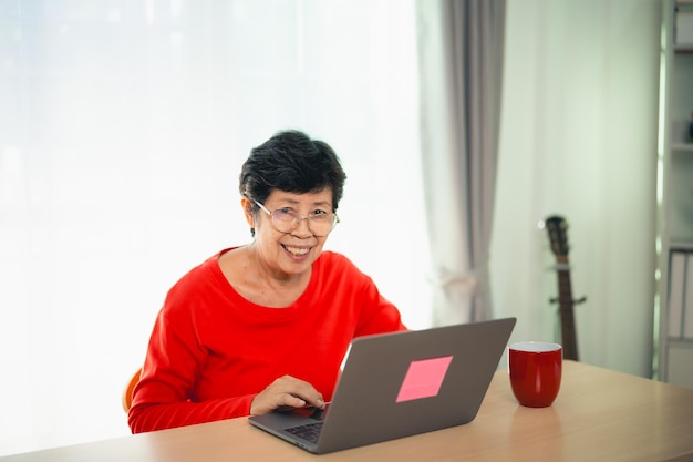 Mujer asiática anciana que trabaja después de jubilarse usando una computadora portátil en casa Viejos freelancers que trabajan o aprenden nueva tecnología en una laptop en la sala de estar Concepto de actividad de jubilación