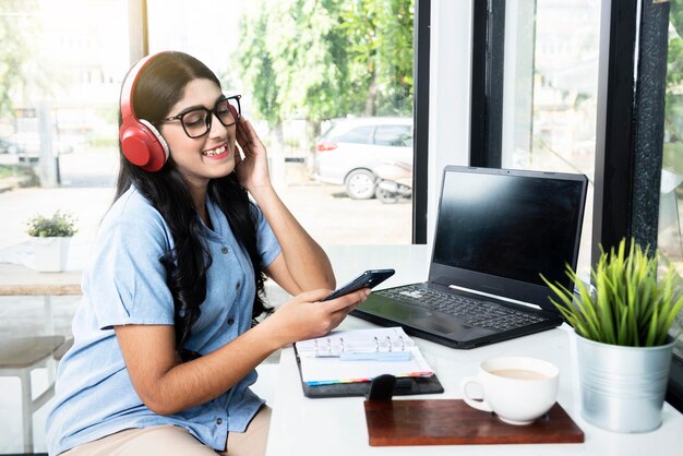 Mujer asiática con anteojos usando un teléfono móvil y auriculares con una computadora portátil y una libreta con una taza de café en la mesa