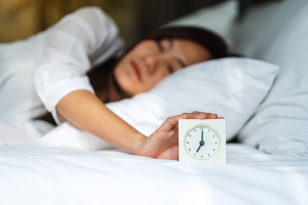 Una mujer asiática apagar un despertador mientras duerme en una cama blanca y acogedora en la mañana