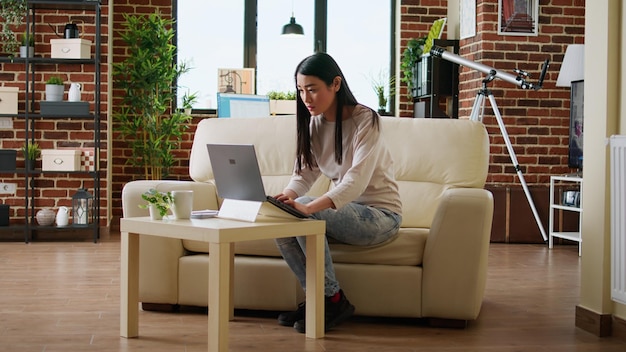 Mujer asiática apresurada que trabaja de forma remota en un proyecto de inicio debido a la fecha límite temprana. Hermosa freelancer haciendo trabajo remoto en una laptop moderna mientras desarrolla ideas de negocios en casa.