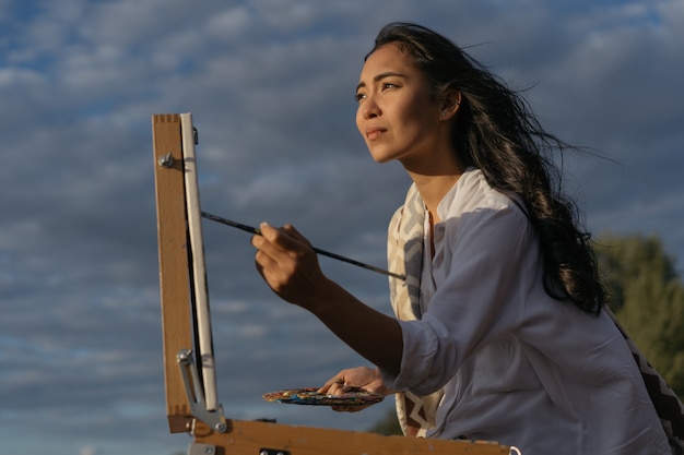 Mujer asiática artista pintando cuadro sobre lienzo, sosteniendo pincel y paleta