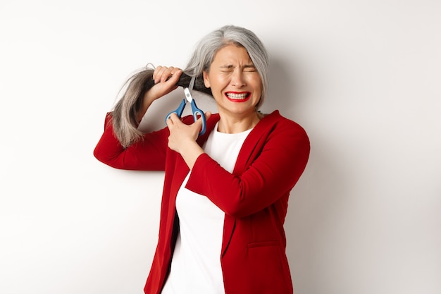 Mujer asiática asustada cortando el pelo con unas tijeras y sintiéndose ansiosa, cierra los ojos con nerviosismo, de pie en una chaqueta roja sobre fondo blanco.