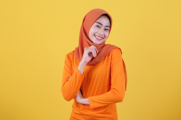 La mujer asiática atractiva alegre demuestra el espacio de la copia en la pared amarilla en blanco, tiene una expresión amistosa feliz, vestida casualmente con el hijab, posa interior