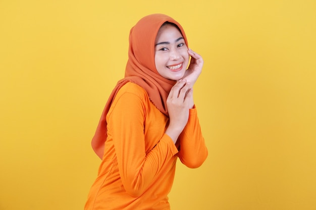 La mujer asiática atractiva alegre demuestra el espacio de la copia en la pared amarilla en blanco, tiene una expresión amistosa feliz, vestida casualmente con el hijab, posa interior