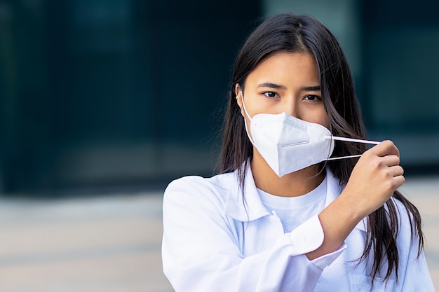 Mujer asiática, atractiva chica étnica en máscara médica en su rostro mirando a cámara con una mirada seria en camisa blanca fuera del centro de negocios