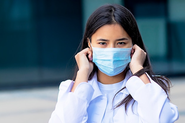 Mujer asiática, atractiva chica étnica con una máscara médica en su rostro, mirando a la cámara con una mirada seria en una camisa blanca fuera del centro de negocios.