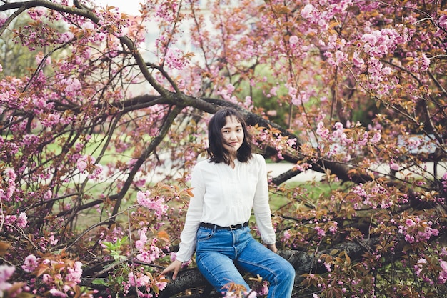 Mujer asiática atractiva joven en una camisa blanca que se sienta bajo el árbol floreciente de sakura en la calma del parque