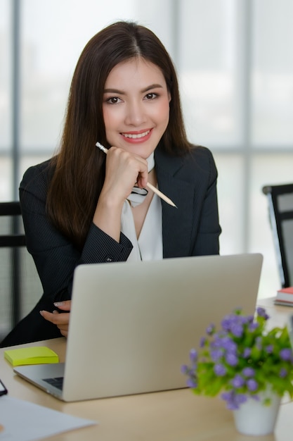 Mujer asiática atractiva joven en negocio negro que se sienta detrás de la mesa con la computadora y el ordenador portátil en la oficina que mira moderna con el fondo borroso de las ventanas. Concepto de estilo de vida de oficina moderna.