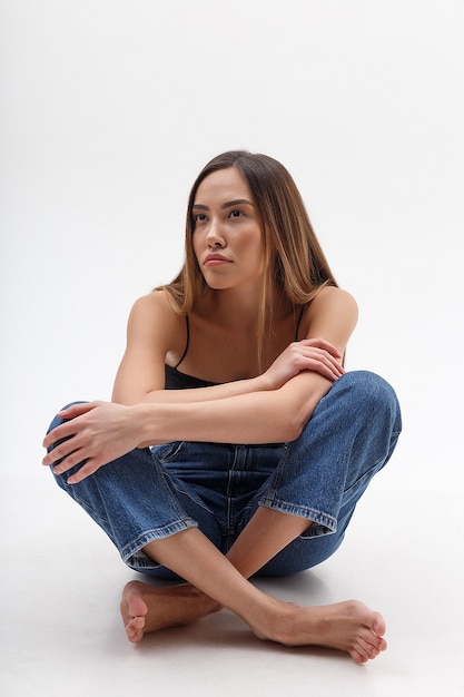 Mujer asiática atractiva joven con el pelo largo en la parte superior negra, jeans azul aislado en la pared blanca. mujer bonita flaca sentada en ciclorama con las piernas cruzadas. pruebas modelo de bella dama