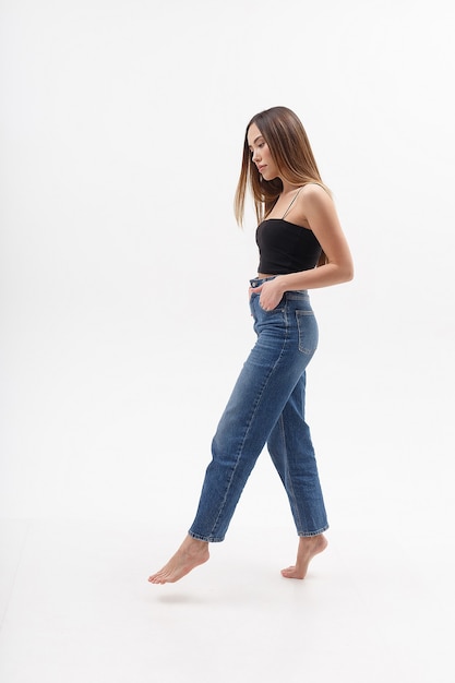 Mujer asiática atractiva joven con el pelo largo en la parte superior negra, jeans azul aislado sobre fondo blanco de estudio. mujer bonita flaca posando en ciclorama con los pies descalzos. pruebas modelo de bella dama