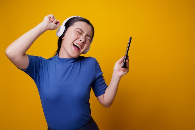 Mujer asiática con auriculares escuchando música desde un teléfono inteligente en amarillo.