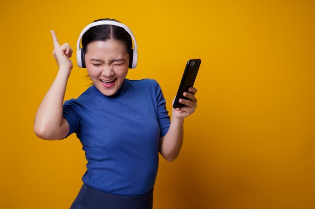 Mujer asiática con auriculares escuchando música desde un teléfono inteligente en amarillo.