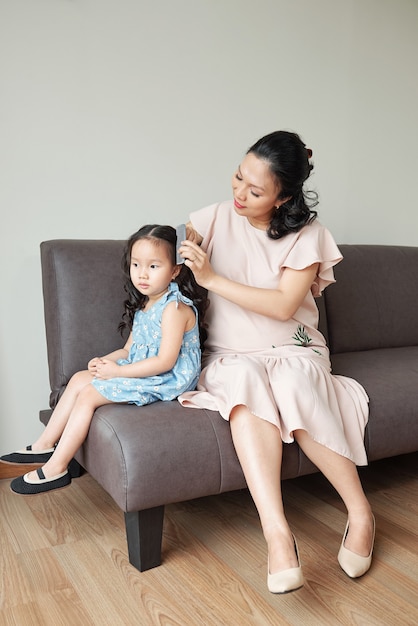 Mujer asiática bastante embarazada sentada en el sofá con su hija y peinando el cabello de su pequeña hija cuando se preparan para la celebración