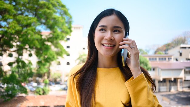 Mujer asiática bastante joven hablando con amigos por teléfono móvil mientras está de pie en el balcón del edificio de oficinas