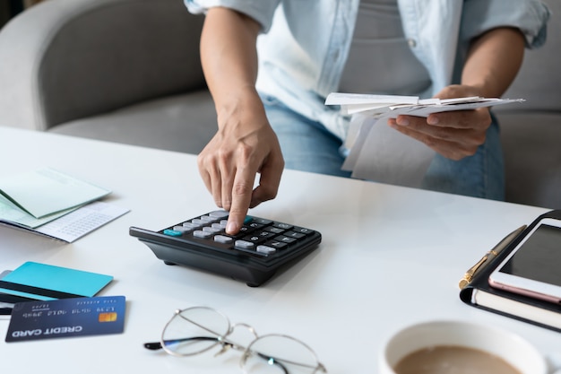 Mujer asiática bastante joven que usa la calculadora mientras que lleva a cabo cuentas para calcular gastos e impuestos caseros en sala de estar en casa.