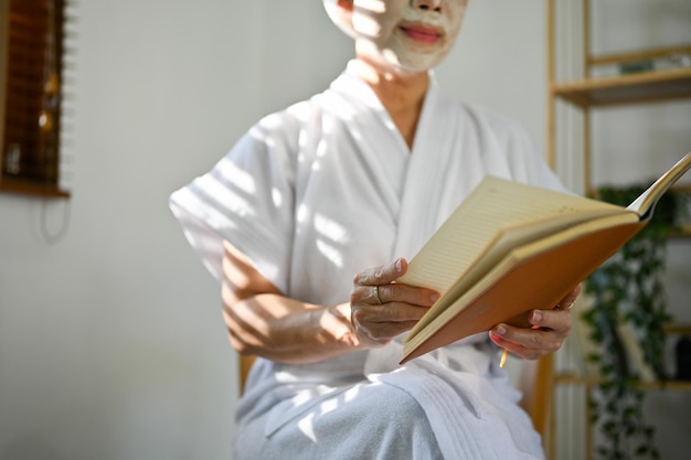 Mujer asiática en bata de baño está leyendo un libro mientras se aplica una máscara de arcilla facial en la imagen recortada de la cara