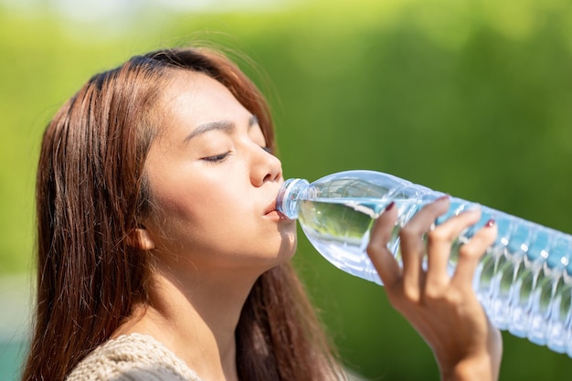 Mujer asiática bebe agua de botella grande en el fondo del árbol verde al aire libre